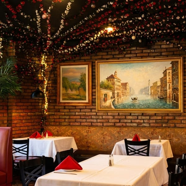 Cozy restaurant interior with brick walls, framed art, red and white floral ceiling decor, and set tables.