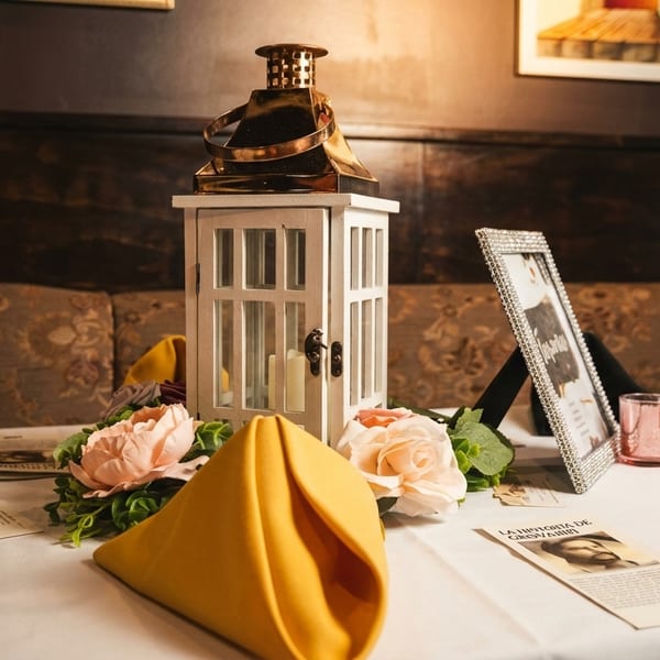 A white lantern centerpiece with candles, pink flowers, and yellow napkins on a white tablecloth.